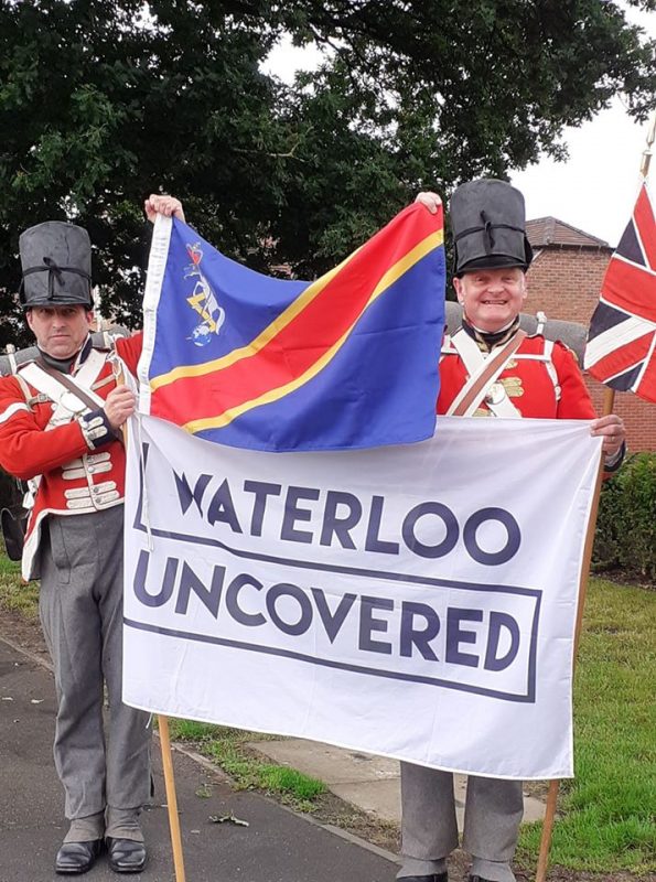 Coldstream Guards reenactors Tony Fielding and Clive Jones pose with a Waterloo Uncovered flag in full Napoleonic British uniform